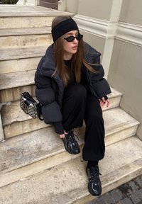 Giacca a vento nera con maniche larghe, pantaloni lunghi neri, sneaker nere e una fascia nera per capelli. Una piccola borsa con motivi si trova sui gradini.