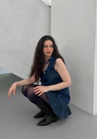 Abito in denim con accenti blu a nastro, collant neri a pois e stivaletti neri alla caviglia. Il modello è accovacciato contro un muro grigio chiaro.