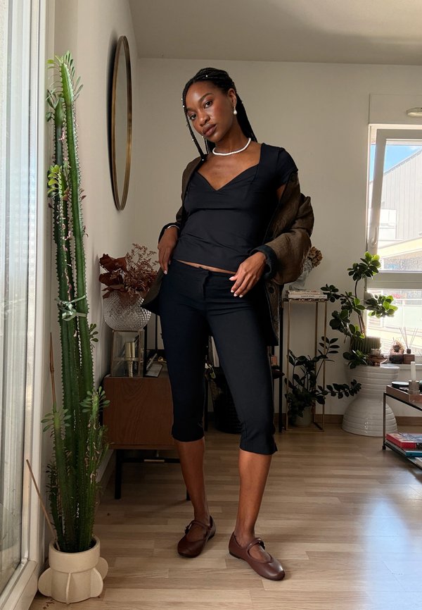 Jonge vrouw met gevlochten haar poseert binnen, gekleed in een zwart topje, korte zwarte broek, bruine schoenen en een bruine jas over haar schouders.