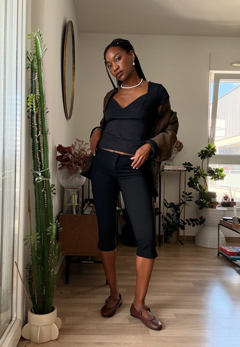 Jeune femme avec les cheveux tressés posant à l'intérieur, portant un haut noir, un pantalon noir court, des chaussures marron et une veste marron sur les épaules.