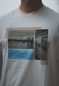 Λευκό βαμβακερό T-shirt με γραφικό σχέδιο που απεικονίζει τέσσερα παραλληλόγραμμα σε διάφορα χρώματα: μπεζ, μαύρο, μπλε και γκρι, με αστική εικονογραφία και κείμενο.