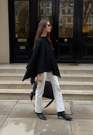 Poncho de lana negro con cuello alto, combinado con vaqueros blancos de pierna recta y botines negros de punta. Incluye un bolso negro.