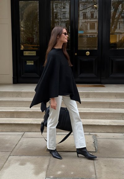 Poncho de lana negro con cuello alto, combinado con vaqueros blancos de pierna recta y botines negros de punta. Incluye un bolso negro.