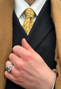 Mano sosteniendo la solapa de un traje negro, con un anillo plateado en forma de calavera, corbata amarilla con patrón de ovejas, camisa blanca y abrigo marrón.