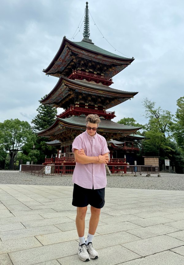 Un hombre con gafas de sol está de pie con los brazos cruzados frente a una ornamentada pagoda japonesa roja y verde, en un patio pavimentado.