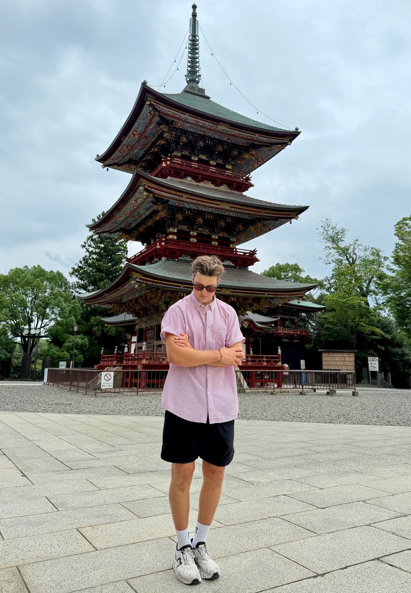 Un hombre con gafas de sol está de pie con los brazos cruzados frente a una ornamentada pagoda japonesa roja y verde, en un patio pavimentado.