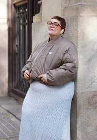 Άτομο με κοντά μαλλιά και μεγάλα γυαλιά ακουμπάει σε πέτρινο τοίχο φορώντας μπεζ μπουφάν και υφασμάτινη ανοιχτό γκρι φούστα σε εξωτερικό χώρο.