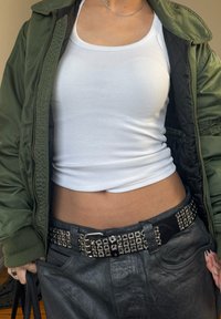 Osoba ubrana w biały krótki top na ramiączkach, zieloną kurtkę bomberkę, ćwiekowany czarny pasek oraz ciemne spodnie, trzyma czarną torebkę.