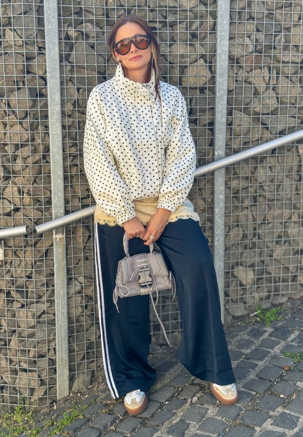 Femme avec des lunettes de soleil portant une veste blanche à pois, un pantalon large bleu marine avec des rayures blanches, un top beige, tenant un petit sac à main gris, debout près d'un mur en pierre.