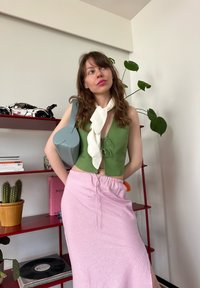 Top verde curto com cordão na frente, lenço branco e saia rosa com cordão. Bolsa azul claro em forma de coração e cacto em vaso numa prateleira vermelha.