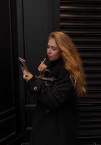 Sort overcoat med knapdetaljer; holder en smartphone i den ene hånd og en læbestift i den anden; tekstureret mørk baggrund. Langt, bølget hår.