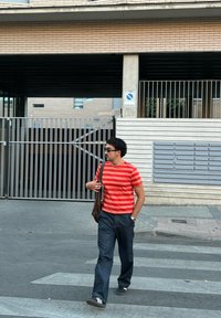 Muž v červeném pruhovaném tričku, tmavých kalhotách a slunečních brýlích kráčí po přechodu a nese hnědou tašku přes rameno v městském prostředí.