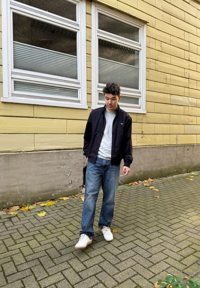 Chaqueta bomber de la marina sobre suéter gris, jeans azules y zapatos blancos. Está de pie sobre un camino de ladrillos con hojas caídas y un fondo de pared amarilla.