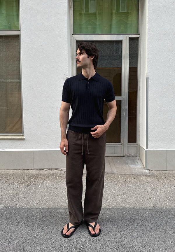 Mann mit dunklem Haar und Schnurrbart, der ein schwarzes Kurzarmshirt, braune Hosen und schwarze Flip-Flops trägt und draußen neben einer Glastür steht.