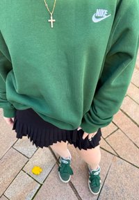 Pessoa a usar um moletom verde da Nike, saia plissada preta, sapatilhas verdes e um colar de cruz dourado, a pé em pavimento de azulejos.