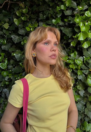 Yellow ribbed T-shirt, pink handbag strap, gold flower earrings with a blue center, long blonde hair, green ivy background.