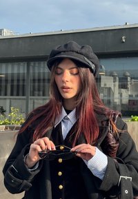 Jeune femme aux longs cheveux roux portant un chapeau noir et un manteau tient des lunettes de soleil noires à l'extérieur dans un environnement urbain avec des bâtiments en arrière-plan.