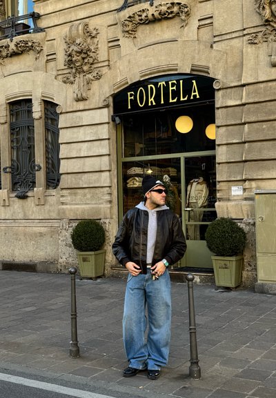 Chaqueta bomber de cuero negra sobre una sudadera gris, combinada con unos vaqueros de pierna ancha en azul claro y zapatos negros. Rodeado de macetas verdes.