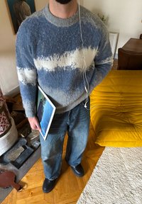 Pull en tricot épais bleu et blanc avec un motif texturé ; jeans bleus et chaussures noires ; tenant un cadre photo.