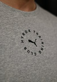 Close-up van een grijs stoffen shirt met een zwart rond logo waarop staat "HYROX TRAINING CLUB" rondom een puma-silhouet.