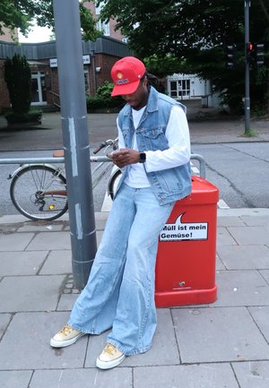 Chaleco de denim sobre una camisa de manga larga blanca, jeans de pierna ancha de color azul claro, zapatillas amarillas, gorra roja con logo, de pie cerca de un cubo de basura.