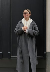 Grå ullfrakk med stor krage, lagvis over en lys genser og et hvitt skjerf. Står mot en svart vegg. Briller på.