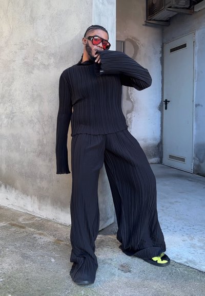Top negro plisado con mangas largas, combinado con pantalones plisados a juego. Presenta un corte relajado y una tela texturizada. Incluye grandes gafas de sol rojas.