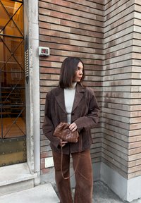 Blazer de pana marrón, cuello alto crema, pantalones de pana marrón. Sosteniendo un bolso marrón pequeño con textura tejida. De pie contra una pared de ladrillos.