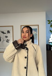 Fleecová bunda krémovej farby s vysokým golierom, čiernymi gombíkmi a kontrastnými švami. Hnedé kožené manžety pridávajú textúrovaný akcent.