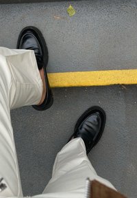 Μαύρο δερμάτινο λουστρίνι με έντονη μύτη και υφασμάτινη σόλα, συνδυασμένο με ανοιχτό μπεζ παντελόνι, πάνω σε γκρι επιφάνεια με κίτρινη γραμμή.