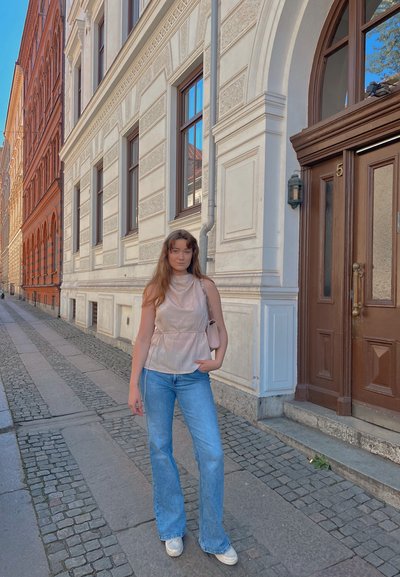 Top sin mangas de color beige claro con un escote único y cordón, combinado con jeans acampanados azules y zapatos blancos. Fondo de una calle pavimentada de piedra.