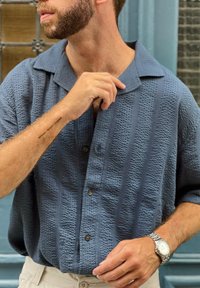 Camisa de manga curta azul marítimo com tecido texturizado e riscas verticais, apresentando um collar camp e cinco botões. Um relógio prateado é visível.