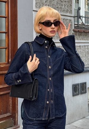 Dunkelblaues Denim-Hemd mit einem Kentkragen, silbernen Knöpfen und einzigartigen Stickmustern. Kombiniert mit einer schwarzen Handtasche und einem schwarzen Rollkragenpullover.