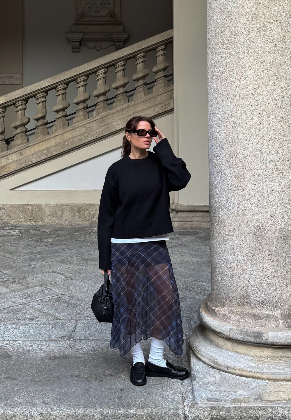 Zwarte oversized trui, doorschijnende geruite rok in marineblauw en wit, gelaagd met een witte top. Zwarte handtas en loafers. De setting bevat stenen kolommen.