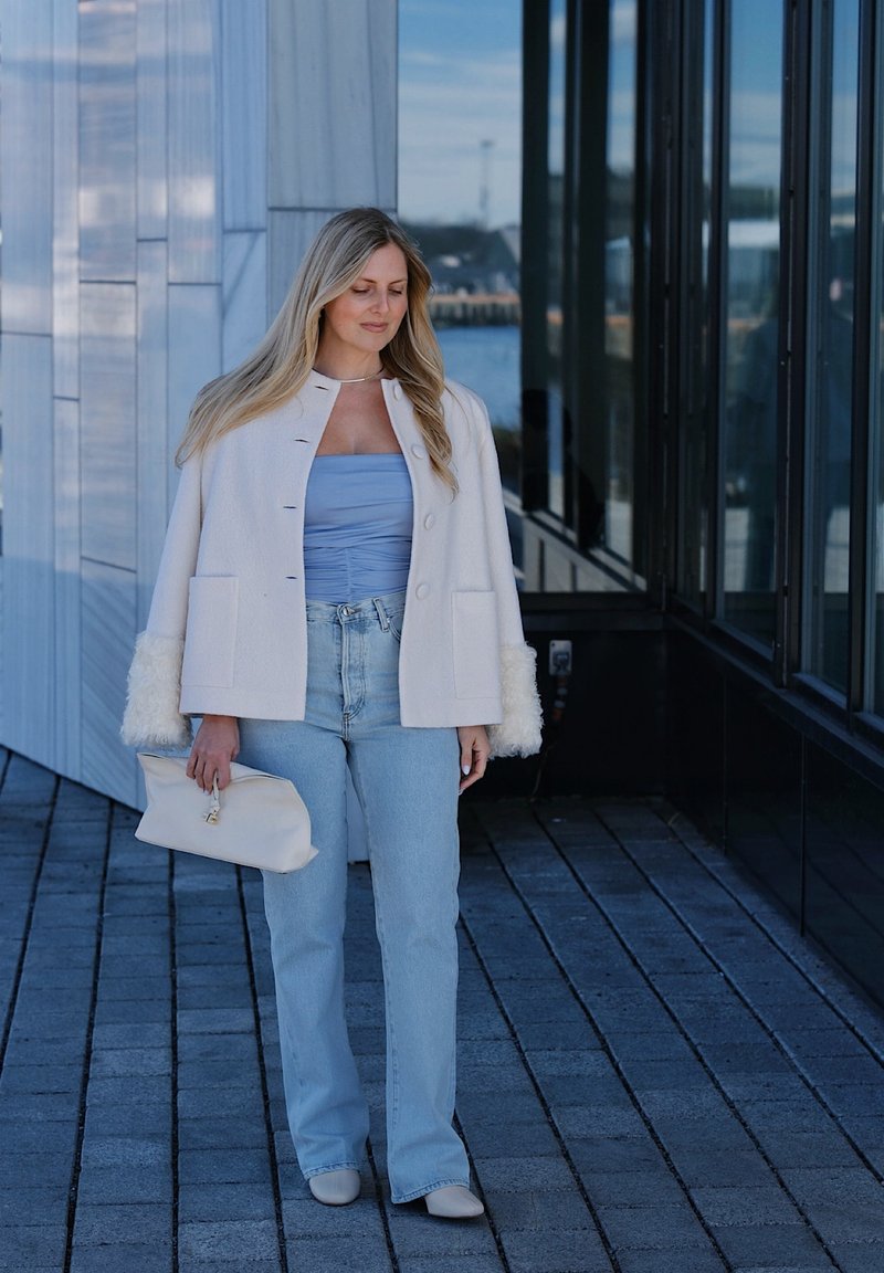Femme en top bleu clair, veste blanche, jean clair et chaussures beige tenant une pochette crème, marchant à côté d'un bâtiment moderne avec des panneaux en verre et en métal.