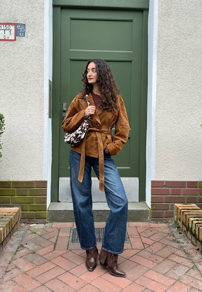 Γυναίκα με σγουρά μαλλιά, που φορά καφέ ζακέτα με ζώνη, μπλε τζιν παντελόνι φαρδιάς γραμμής, καφέ μπότες και κρατάει μια τσάντα με σχέδιο, στέκεται δίπλα σε πράσινη πόρτα.
