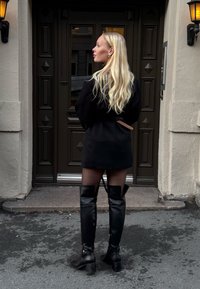 Must kohandatud kleit vööga, läbipaistvad mustad sukkpüksid ja põlveni ulatuvad mustad nahksaapad. Taustal on dekoratiivne uks.