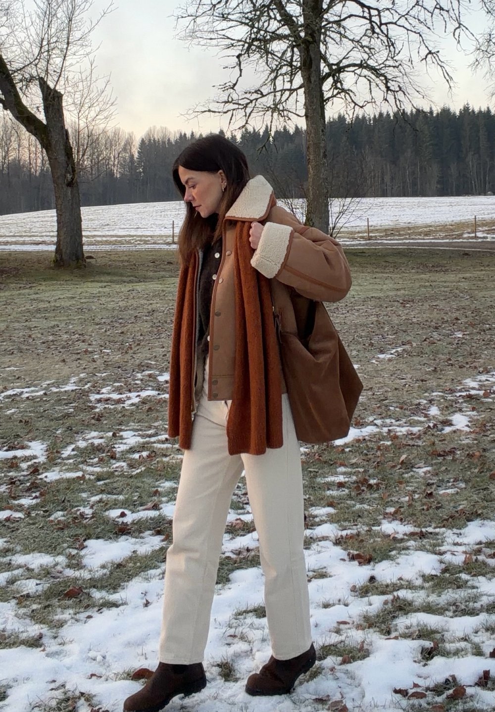 Femme debout à l'extérieur sur une pelouse partiellement enneigée, portant une veste en peau retournée marron, une écharpe rouille, un pantalon crème et portant un sac marron sur l'épaule.