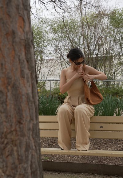 Mujer con atuendo beige sentada en un banco del parque, mirando dentro de su bolso marrón, con árboles y plantas de fondo.