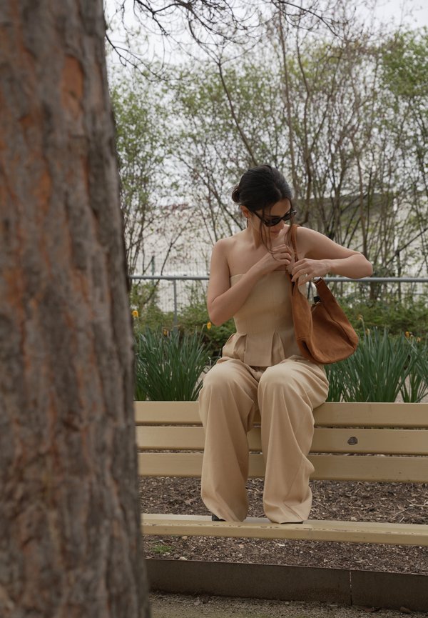 Donna in abito beige seduta su una panchina nel parco, che guarda dentro la sua borsa marrone, con alberi e piante sullo sfondo.