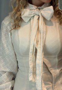 Blusa bianca con un grande fiocco al colletto. Presenta un tessuto strutturato con un delicato motivo geometrico e maniche lunghe a sbuffo.