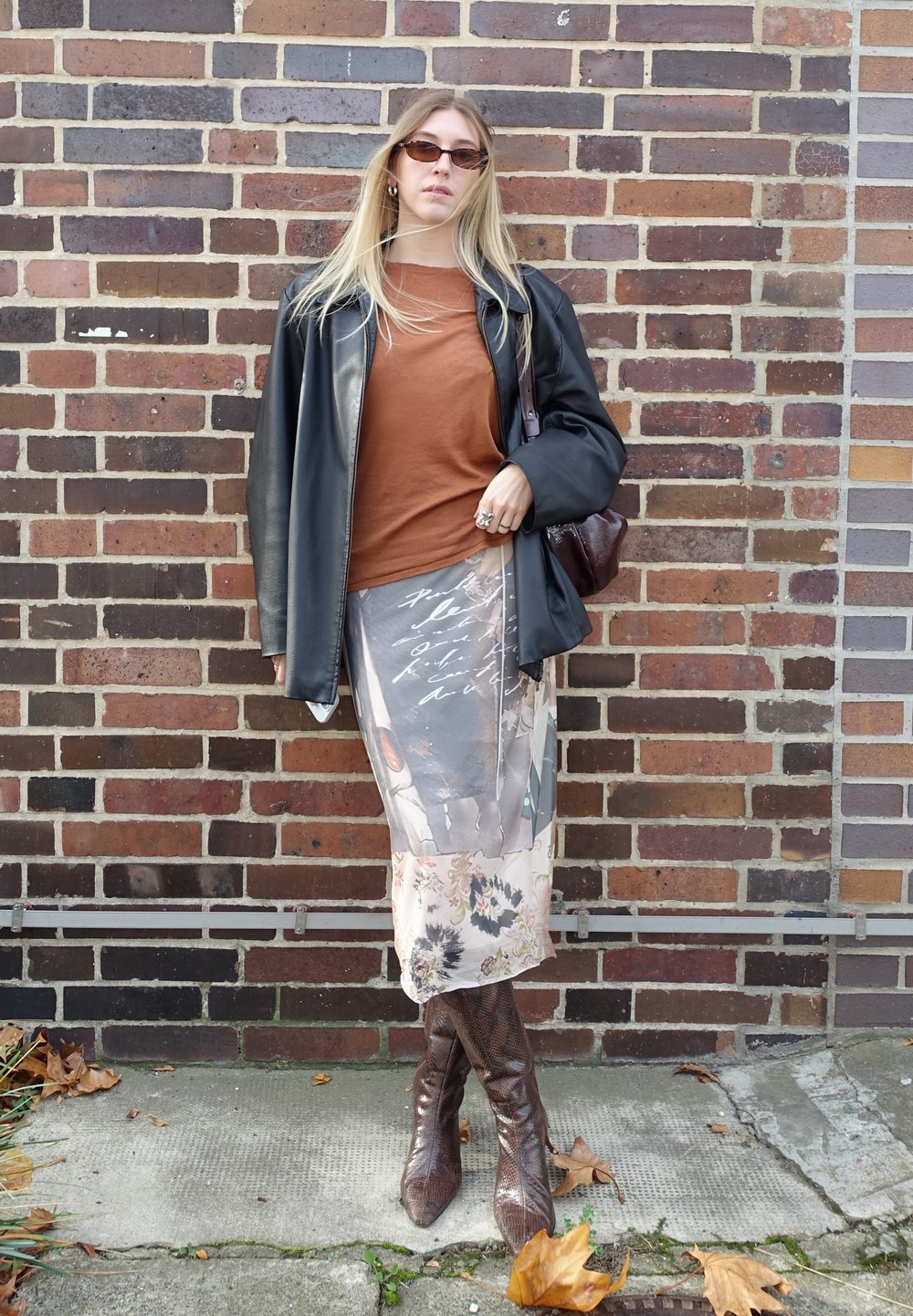 Clear, long black leather jacket over a brown top, paired with a sheer floral-print skirt and brown snake-skin knee-high boots.