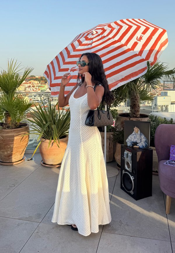 Mujer con vestido blanco y gafas de sol ajustándose el pelo bajo una sombrilla a rayas rojas y blancas en una azotea con plantas en macetas y vistas a la ciudad.