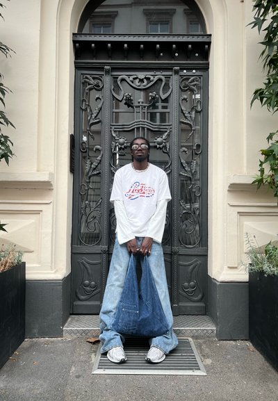 Pantalones de mezclilla en azul claro, diseño de pierna ancha, con un bolsillo trasero. Camisa blanca de manga larga con estampado gráfico colorido. Gafas oscuras.