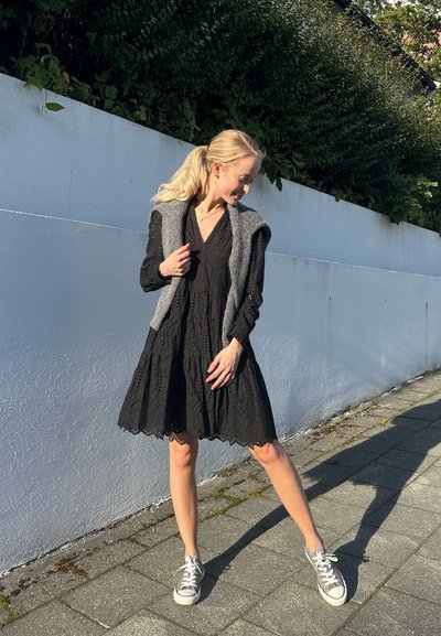 Mujer con coleta rubia, vistiendo un vestido negro de ojales, con un suéter gris colgado sobre los hombros y zapatillas, de pie en una acera soleada junto a una pared blanca.