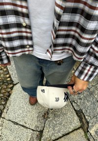 Cappello bianco e nero con logo NY tenuto in mano, camicia a quadri nei colori marrone, blu navy e bianco, jeans azzurri chiari, scarpe marroni. Pietre testurizzate sotto.