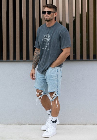 Camiseta gráfica gris, pantalones cortos de mezclilla azul claro desgastados con dobladillo deshilachado, zapatillas blancas y gafas de sol negras. De pie contra una pared clara.