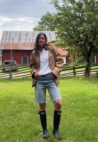 Chaqueta impermeable marrón, top de punto trenzado blanco, pantalones cortos de mezclilla desgastados y botas de goma negras hasta la rodilla en un entorno al aire libre con hierba.