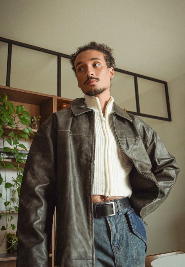 Veste en cuir marron à col montant, portée sur un pull en maille crème. Tissu texturé et coupe ajustée. Jean bleu, ceinture noire.