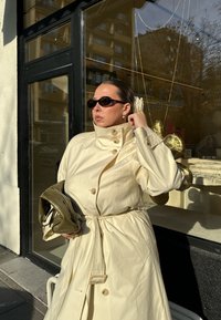 Krém színű ballonkabát magas gallérral, gombos részletekkel és övvel a deréknál; zöld kézitáskával és sötét napszemüveggel párosítva.